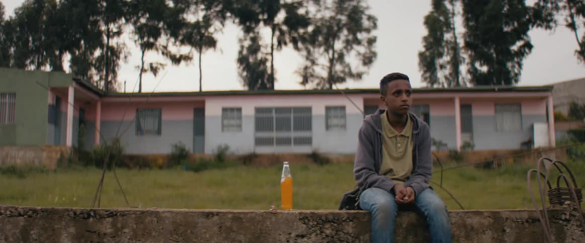 A young man sitting on a short wall next to a boy pushes himself off the wall and walks away with a backpack