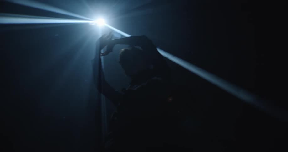 A woman stands under a spotlight with her arms raised above her head