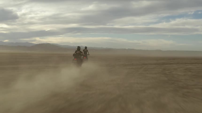 Two motorcyclists kicking up dust while riding across a dirt field on a cloudy day