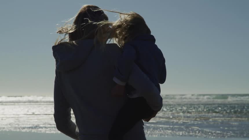 A woman stands by the ocean hold a young girl and their hair blowing in the wind