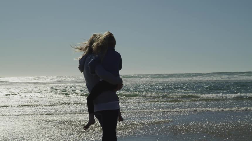 A mother holds her daughter while standing on the beach watching the ocean waves