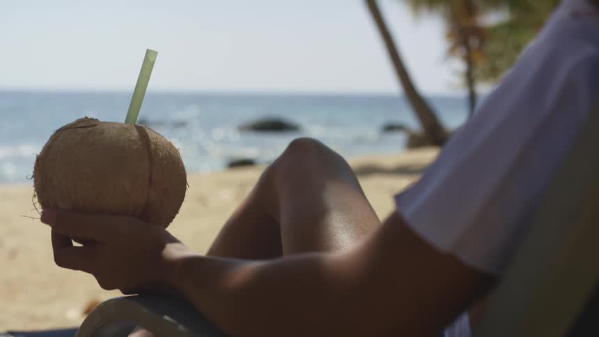 A woman reclines in a chair on a beach as she holds a coconut with a straw in it