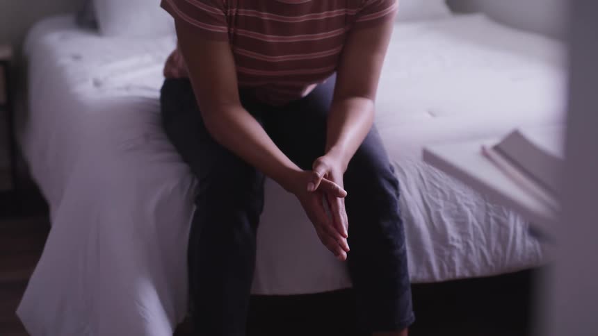 A woman sits on the foot of her bed hunched over with her hands clasped