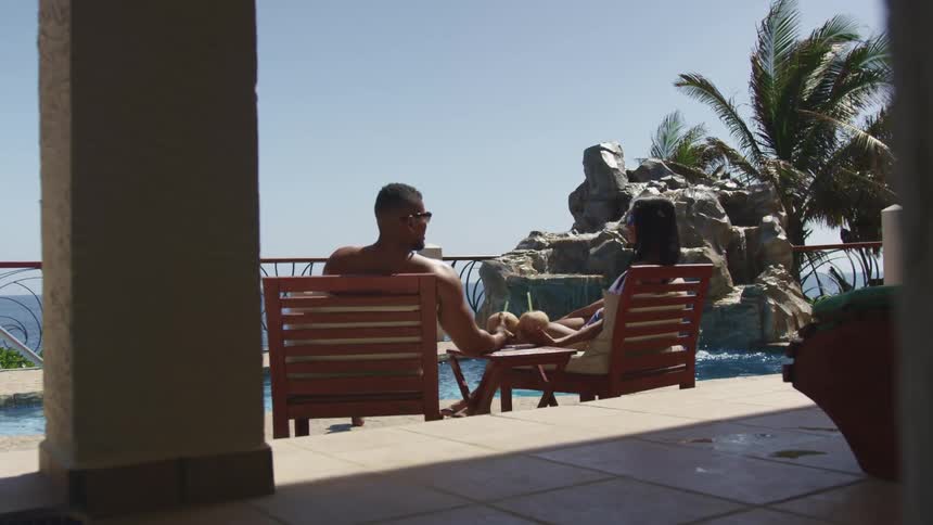 Man and a woman sitting on wooden lounge chairs on a patio drinking from coconuts in front of a pool