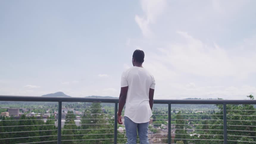 A man stands on his balcony and looks over the neighborhood below to see mountains in the distance