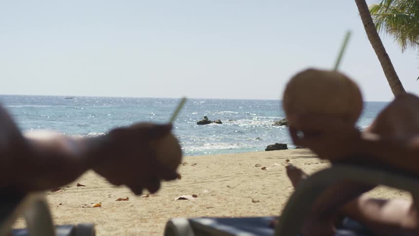 A man and woman lie on loungers on the beach holding drinks in coconuts