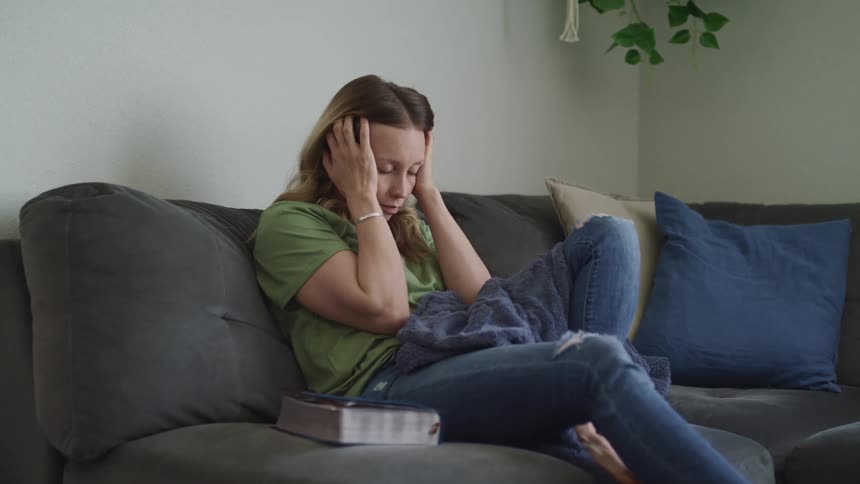 A woman reading a thick book lays it on the sofa beside her before anxiously running her hands through her hair and resting with her eyes closed