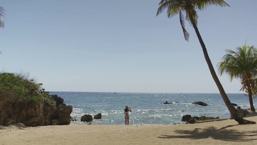 Young woman wearing a swimsuit and sun hat stands on the sand of a tropical beach