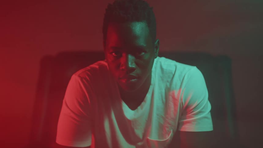A black teenager male wears a white t shirt sitting indoors in shadows with a red hue