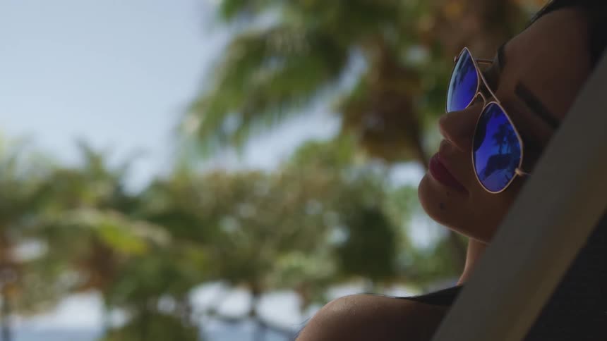 Woman sits in chair in shade on sunny day with sunglasses on and palm trees in the background