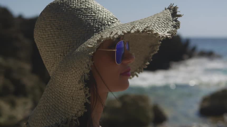 Young woman wearing sunglasses and a sun hat standing on the coast of a beach
