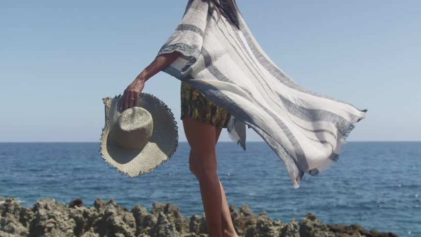 A woman holding a hat near a coast with the ocean sky and clouds in the background