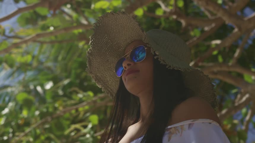 A woman stands under a large tree outside wearing a straw hat and sunglasses