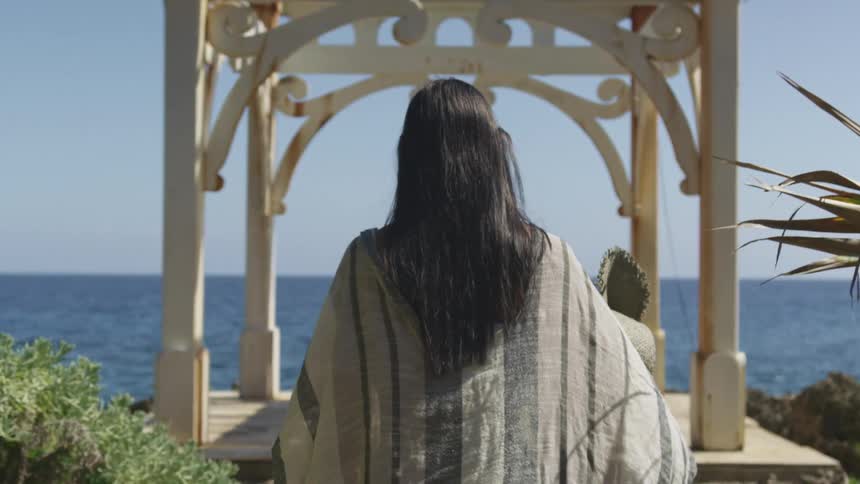 A woman slowly walks towards a platform and pergola that overlook the ocean on a sunny day