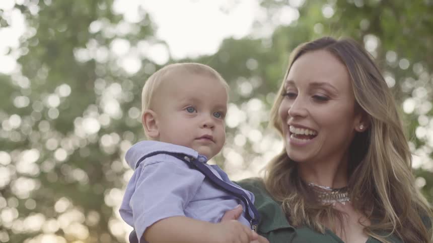 A woman in a green dress holding a baby boy with trees the sky and clouds in the background