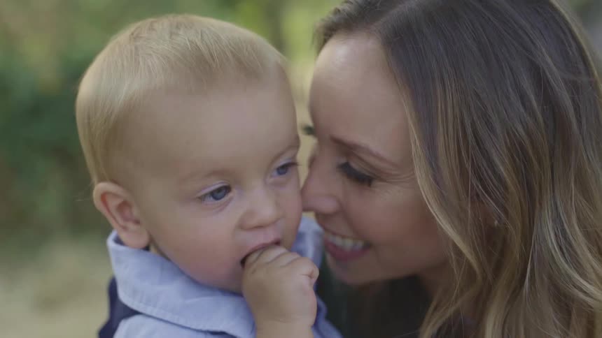 A woman smiles and nuzzles her baby boys cheek as they stand on a lawn