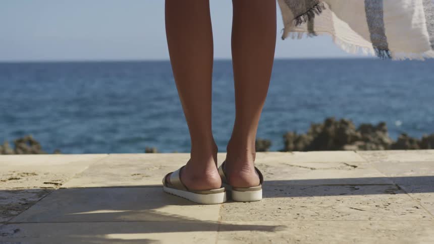 A woman in sandals stands outdoors on a sunny day nearby to the blue ocean