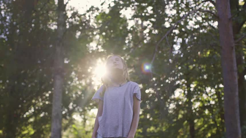 A young girl stands in a forest and looks up towards the sky and closes her eyes