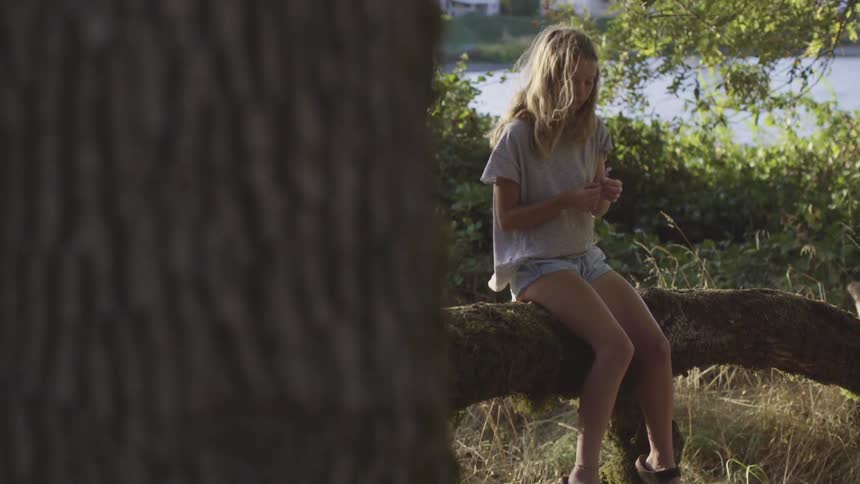Blonde girl sitting on the fallen branch of a tree near a river