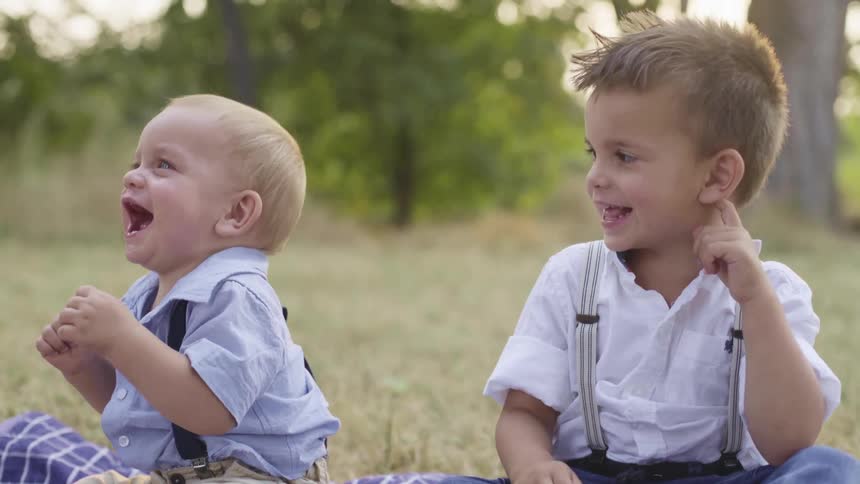 Two little boys sit on a blanket on the grass and laugh