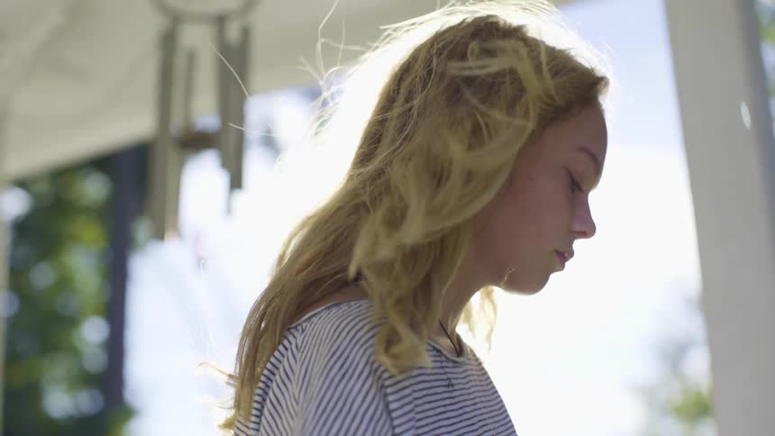 Young girl standing on a patio with wind chimes hanging overhead while she looks down sadly