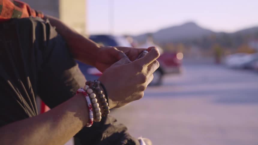 A man sits his knee pulled up and scrolls through his smartphone in a parking lot