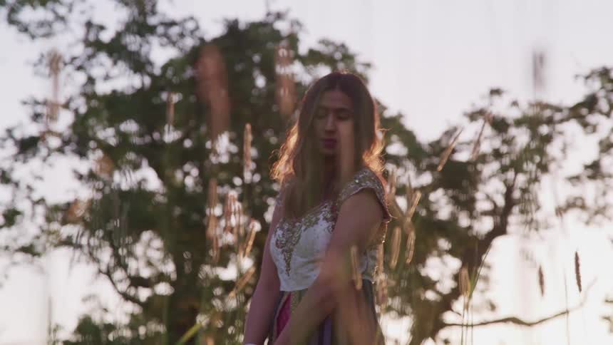 A woman with long hair stands in a field in front of a tree with a blue sky behind her