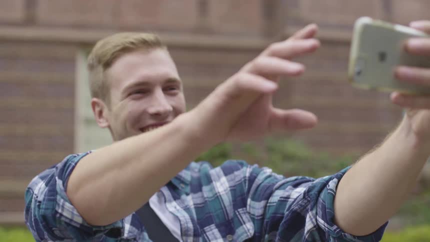 A male teenager wearing a blue plaid shirt takes a selfie while standing outdoors