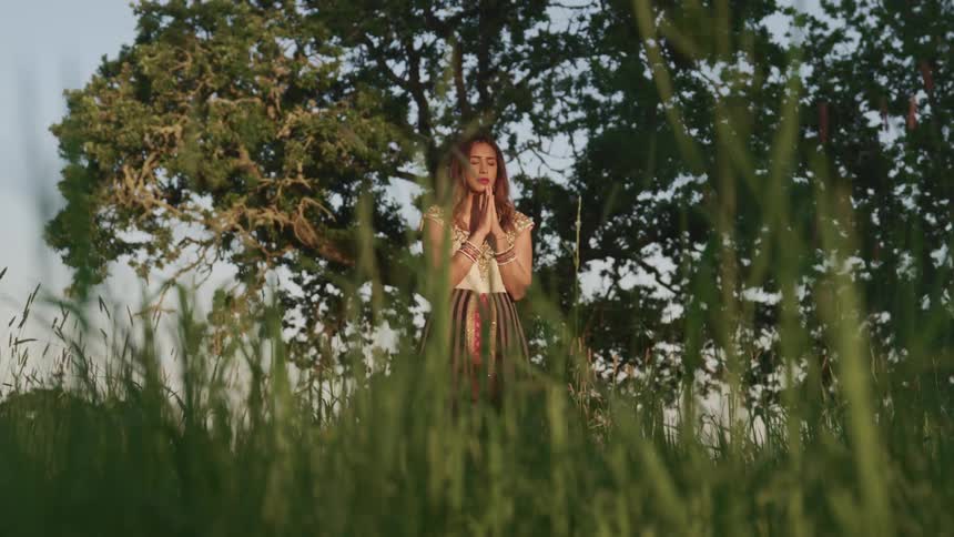 A woman holds her hands together praying while standing outdoors in grass and trees during the day