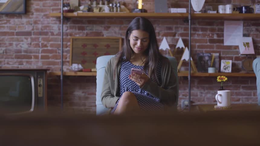 A woman smiles while scrolling through her phone as she sits in a chair in her brick walled apartment