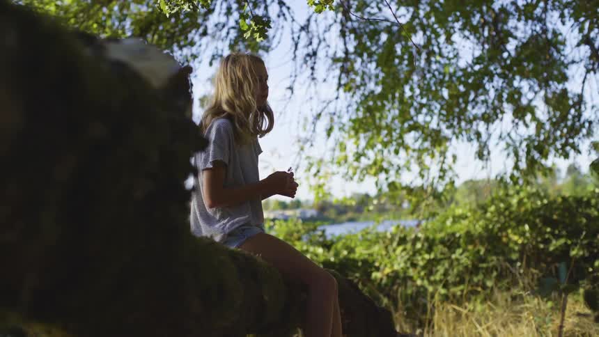 A young girl sits on a fallen tree trunk while in the shade of another tree