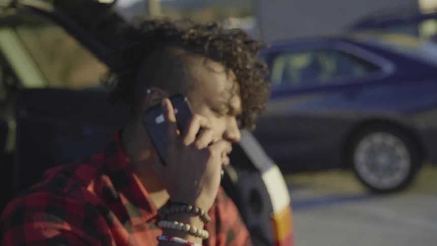 A man sits on the back of a car in a parking lot talking on a phone smiling and laughing