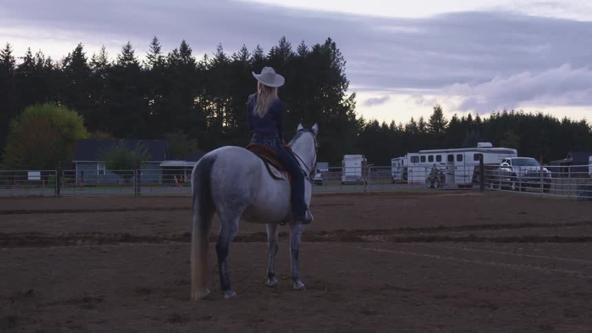 Woman sits atop a white horse on a saddle at a ranch and looks around her in the evening