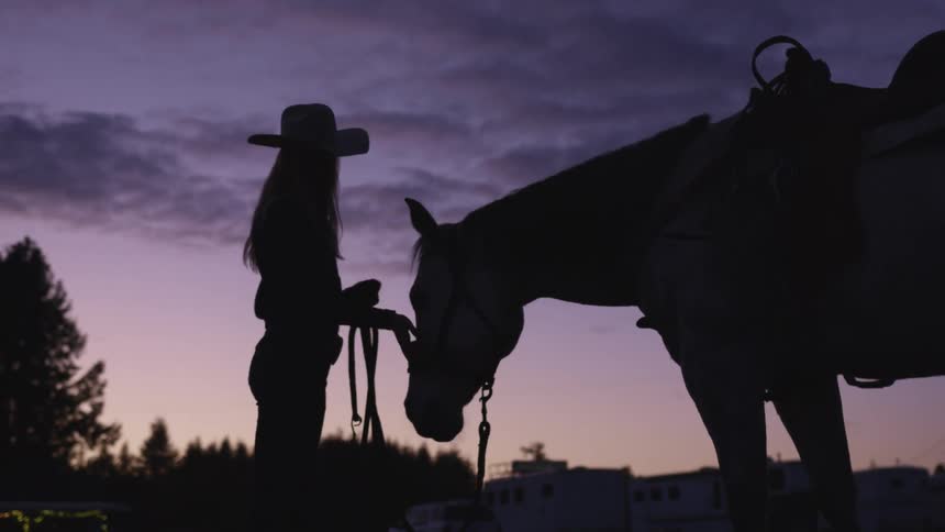 A woman wearing a cowboy hat pets a horse with one hand while holding the reins with the other at sunset