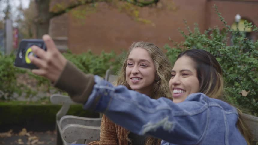 Two girls smiling at the camera on a smartphone taking selfies sitting on a bench