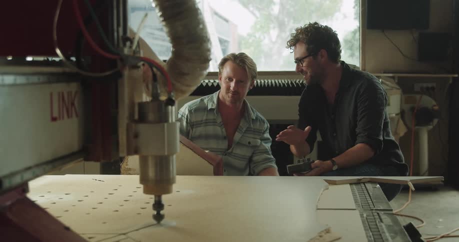 Two men sit inside looking at a drill on a table talking