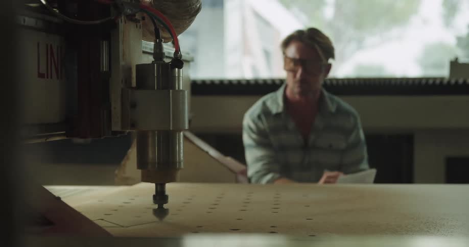 A man sits indoors wearing safety glasses as he writes on paper and looks at a machine on a table