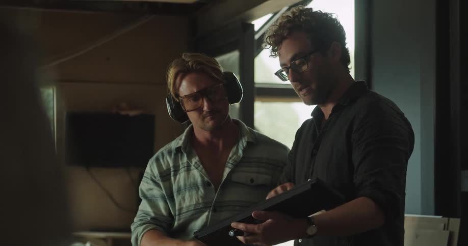 A pair of men standing in a workshop during the day speak while gesturing at a wireless keyboard