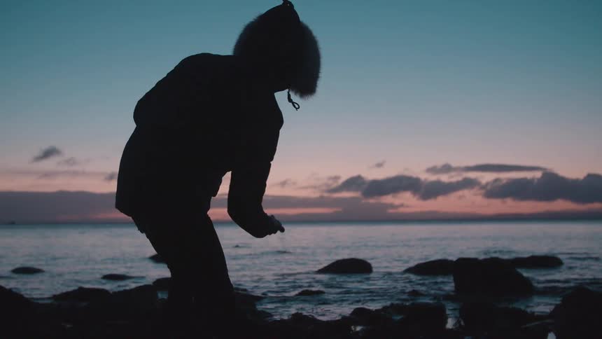 A person picks up a stone and skips it into the ocean at sunset