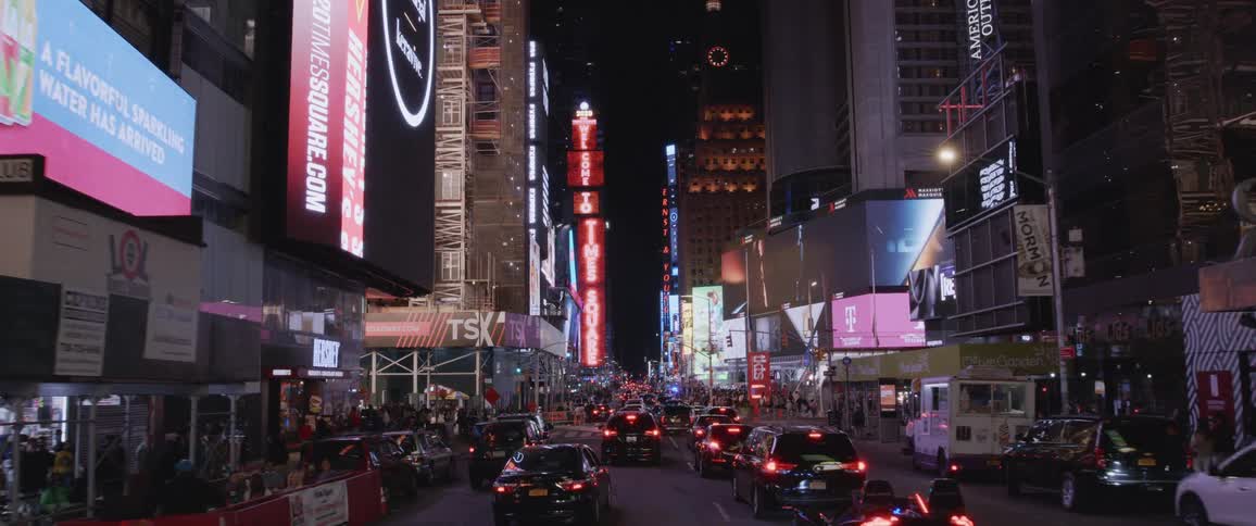 A busy city street with lit up shops and offices and pedestrians walking the streets with cars driving
