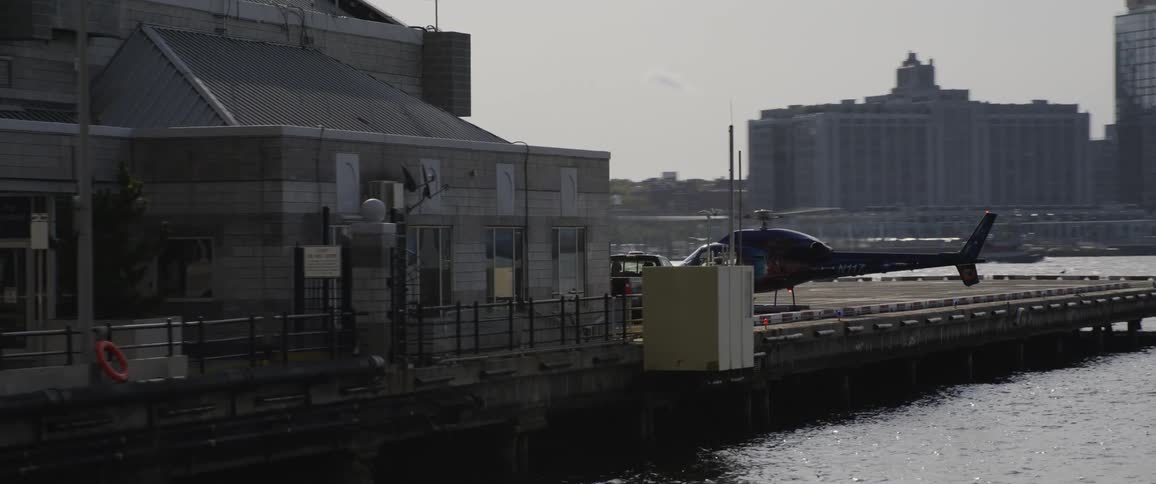 A helicopter parked on a pier as the pilot runs towards a building and people run across a bridge