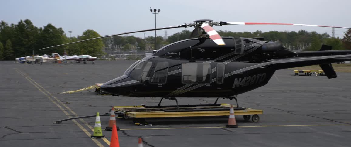 A black helicopter sits on asphalt with other parked airplanes surrounded by trees