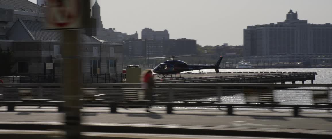 Helicopter lands on landing pad next to a building with a city in the background and people walking on the path in front of the water