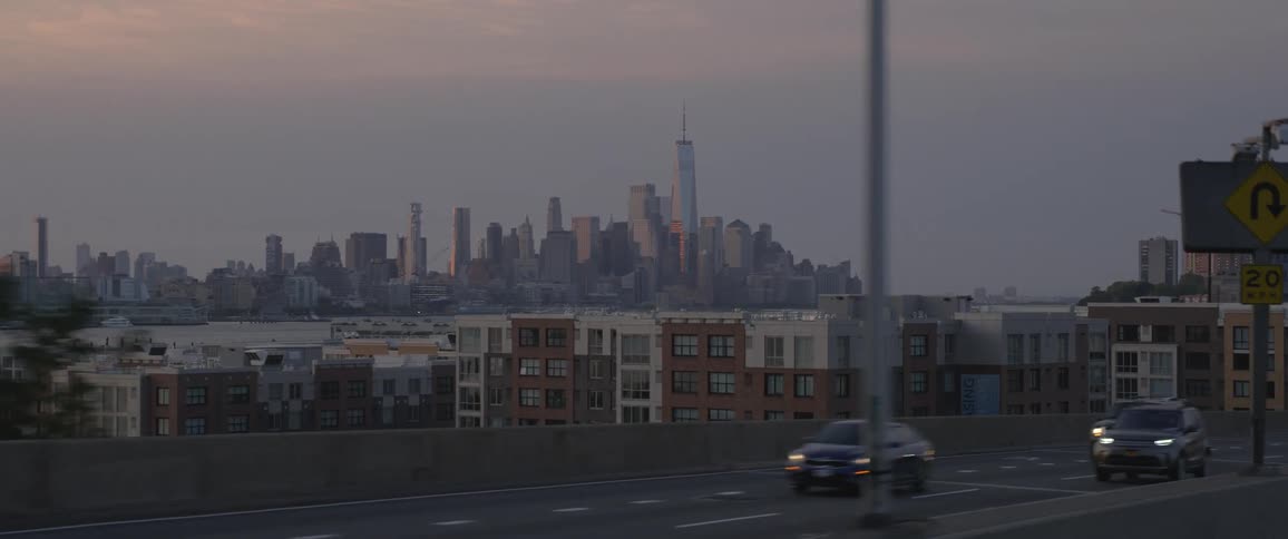 Traffic moves along a highway as the skyscrapers of a metropolis loom in the distance across a bay in the evening