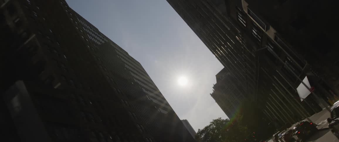 Sun flares as it shines it between rows of tall buildings with cars parked on the road below them