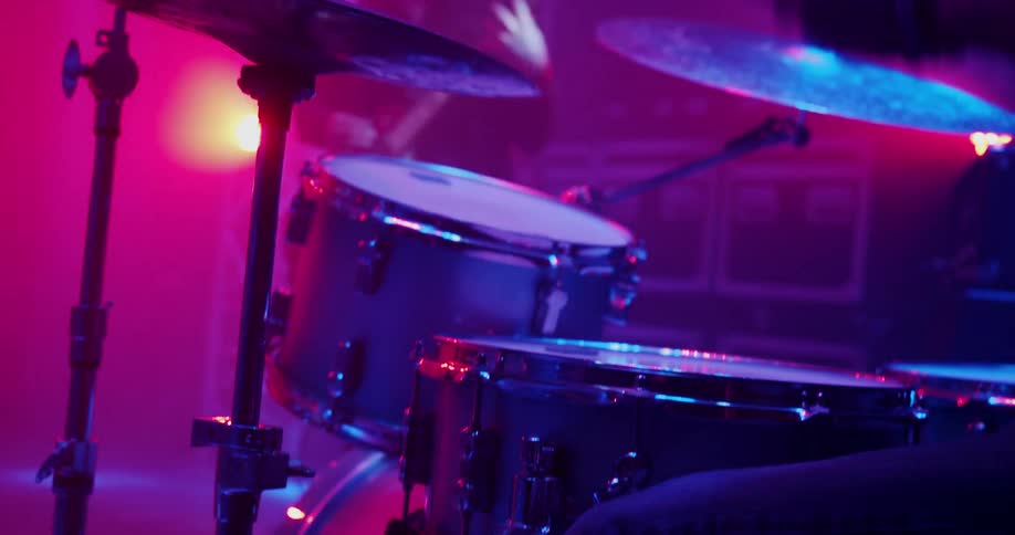 Man plays drums under colored lights on stage with man playing guitar in the background