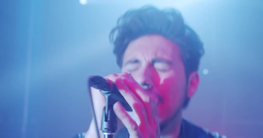 A man with a beard and wearing a leather jacket signs on stage with a microphone with lights flashing
