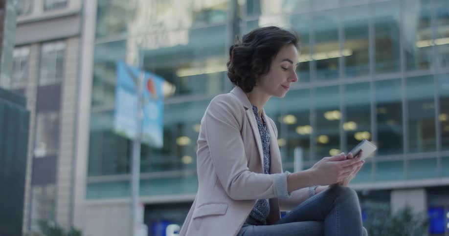 A woman sits and swipes on a tablet in front of a window downtown