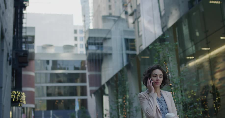 A young woman holding a disposible coffee cup talks on a smartphone while standing outdoors in a city during the day