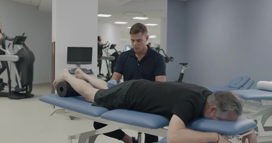 People work on machines at a physical therapy clinic as a man lies face down while another man treats his injury